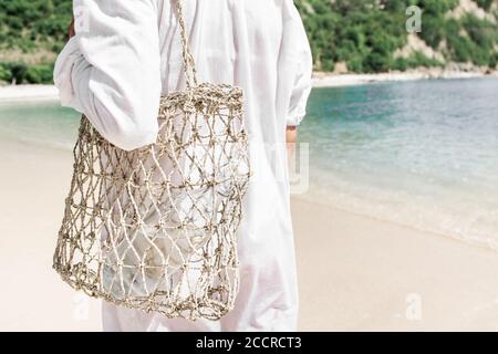 Woman holding straw wicker reusable bag with glass bottle for water. Plastic free and zero waste lifestyle. White sand beach with clear turquoise water on background. Stock Photo