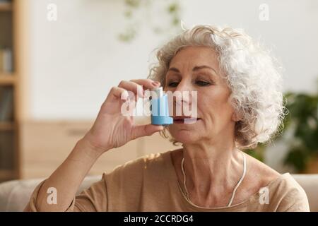 Warm toned head and shoulders portrait portrait of senior woman using inhaler for asthma or breathing problems in home setting Stock Photo