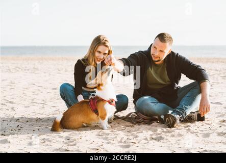 Young happy couple of man and woman with corgi dog siting at sand. Two persons, male feeding pet enjoying beautiful sunny afternoon on beach Stock Photo
