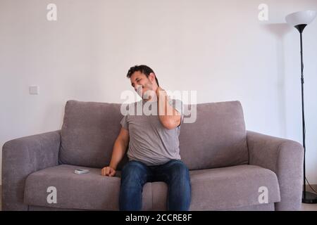 Young man sitting on a sofa suffering from neck pain. Neck and shoulder paint. Stock Photo
