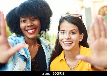 Two beautiful womans of different nationalities show the photo frame with their hands and smile at the camera. Young girlfriends take a selfie on a city street. Stock Photo