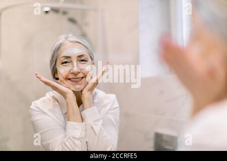 Pretty senior woman with gray hair, wearing white shirt, looking at her face in the bathroom mirror, and applying anti-wrinkle cream or cosmetic mask Stock Photo