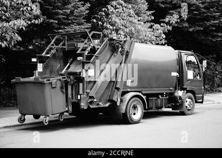 Black and white monochrome image green garbage truck vehicle with an elevated wheelie bin at the rear which is collecting dustbin rubbish Stock Photo