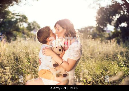 Beautiful, young mom kissing her son on the lips. Boy holding a rose for his mom. Outdoor, nature photo. Mothers day Stock Photo