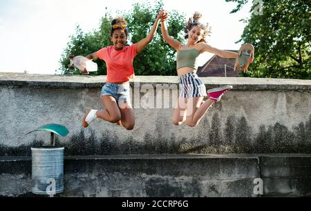 Young teenager girls friends with skateboards outdoors in city, jumping.