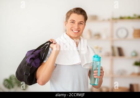 Smiling man with towel, bottle of water and sports bag in hands in interior Stock Photo