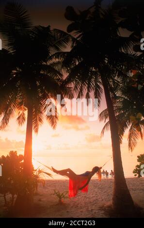 Woman in hammock between palm trees on tropical island holiday silhouetted in sunset light