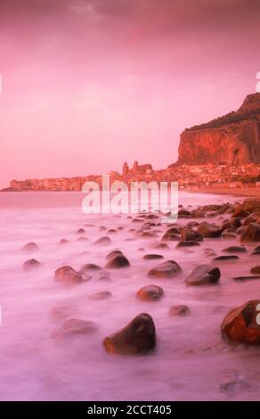 Waves painting rocky shore with Duomo Cathedral above Mediterranean village of Cefalu in Sicily at dusk Stock Photo