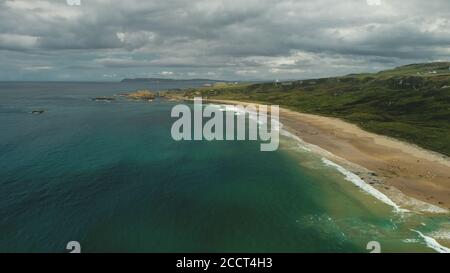Ireland sand beach aerial view: ocean waves, sandy coastline, white shore with greenery meadows. Epic Irish landscape with gray clouds on sky in summer day cinematic shot