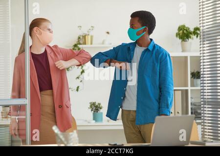 Portrait of young woman wearing mask bumping elbows with African-American colleague as contactless greeting while working in post pandemic office Stock Photo