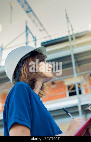 Woman, female engineer, caucasian, age 40, wearing a safety white cap, working on a costruction site in a typical men's role. Gender gap symbol. Verti Stock Photo
