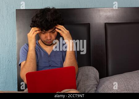 Young Hispanic man working stressed and furious from home on a bed due to quarantine Stock Photo
