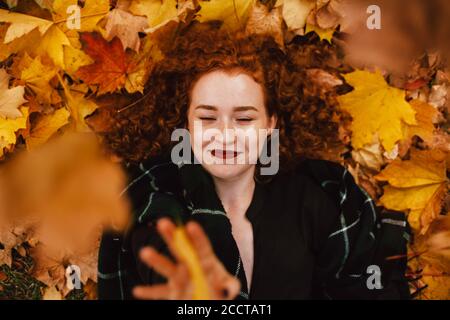 Young happy redhead woman lying on field with orange dry leaves in autumn Stock Photo