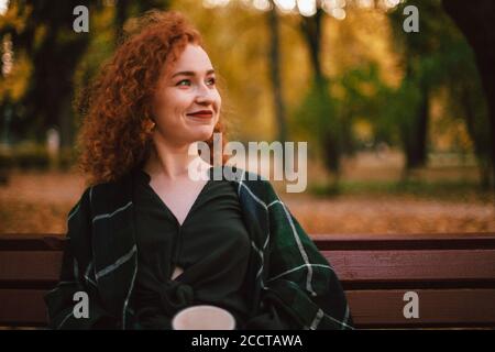 Portrait of smiling red haired woman sitting on bench in park during autumn Stock Photo
