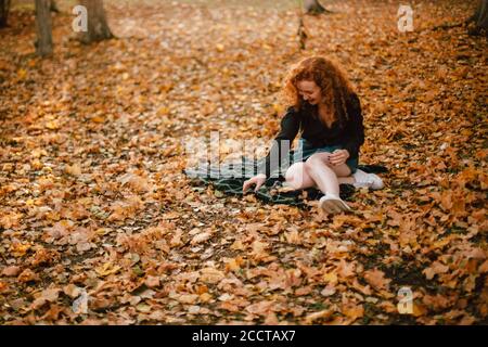 Happy young woman sitting on field with dry leaves during autumn Stock Photo