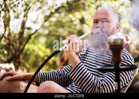 Bearded man smoking hookah outdoors. Hipster dressed in striped sailor shirt exhaling smoke. Relaxation, harmony and tranquility. Camping Stock Photo