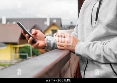 Man drinking coffee or tea on the balcony on the background of mountains and villas or pensions, Close up mans hand using his smartphone. Concept of Stock Photo