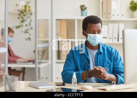 Portrait of young African-American man wearing face mask and sanitizing hands while working in cubicle at post pandemic office, copy space Stock Photo