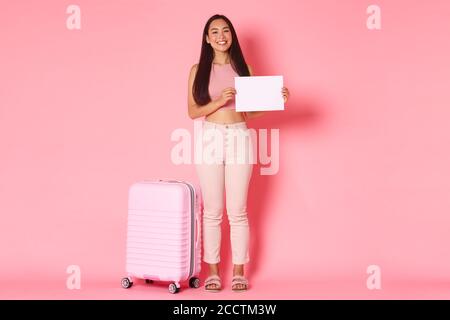 Travelling, holidays and vacation concept. Full-length of beautiful smiling asian girl tourist with suitcase over pink background, waiting for someone Stock Photo