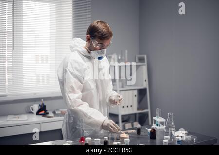 A scientist in protective eyewear, respirator and white overalls standing at the table in the science laboratory, working on something. Stock Photo