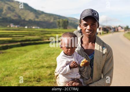 Manandoana, Madagascar - April 26, 2019: Unknown Malagasy man holding child with piece of food on his hand, rice fields in background on sunny day. Pe Stock Photo