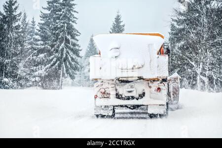 Orange highway maintenance truck removing snow from completely white road during blizzard snowstorm, trees on both sides of road Stock Photo