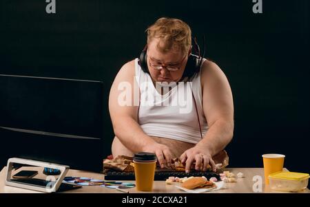 Young obese man in casual clothes sitting at the table and and typing something. close up portrait, black background. lifestyle, weekend Stock Photo