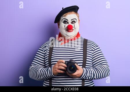 smiling funny clown in stiped sweater holding camera, isolated blue background, studio shot,free time, spare time, leisure Stock Photo