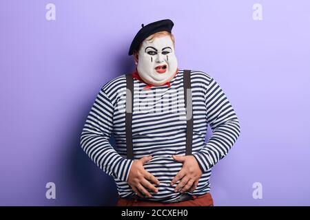 crazy funny plump clown having a problem with his stomach. close up portrait, isolated blue background, studio shot. Stock Photo