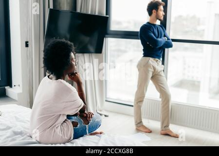 Indoor shot of dark-skinned woman feeling lonely and depressed while her caucasian boyfriend standing next to panoramic window and looking outside. Re Stock Photo