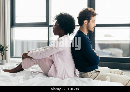 Two unhappy mixed race lovers not talking to each other after having quarrel during holidays at home, sitting on bed and ignoring each other Stock Photo
