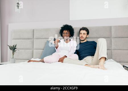 Happy interracial couple in home wear, relaxing in bed holding remote control and watching television Stock Photo