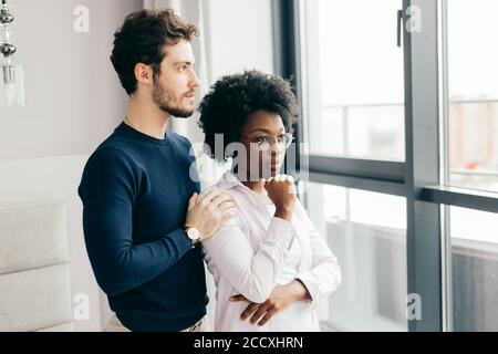 Positive african woman embraces with her european handsome brunette boyfriend, relaxing at home, give warm hug to each other, stand near window backgr Stock Photo