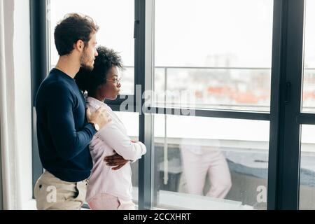 Positive african woman embraces with her european handsome brunette boyfriend, relaxing at home, give warm hug to each other, stand near window backgr Stock Photo