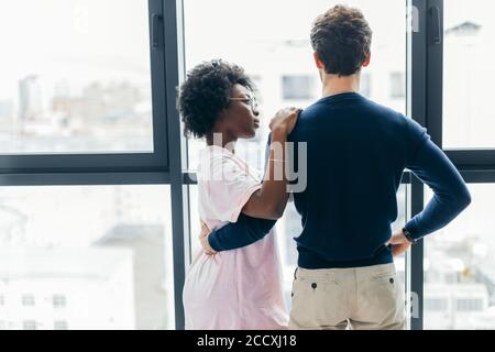 Backside view of young mixed race lovers staying in hotel, Hispanic man hugging African Woman while resting in modern apartment with big window interi Stock Photo