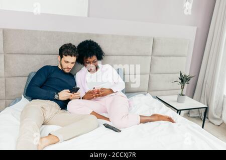 Young wife and husband relaxing on bed during their day-off. Spauses of different nationalities sharing funny stories and watching videos using smart Stock Photo