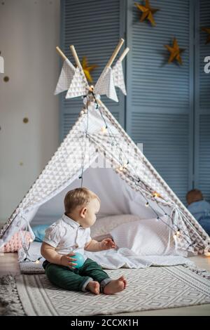 Adorable infant boy sitting on carpet and playing with little children s tent in cozy blue playroom. Toys for little kids. Interior for little boy nur Stock Photo