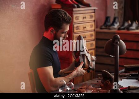 shoemaker in black T-shirt is working with a hammer at workshop, side view photo Stock Photo
