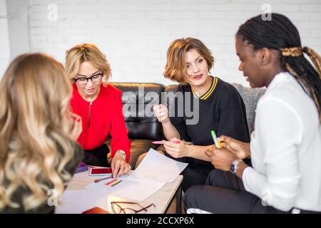 Group of international MLM partners discuss their work in co-working space talking and smiling while sitting at the desk table with smartphones and dr Stock Photo