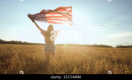 Young woman waves an american flag on the wheat field. Patriotic holiday, independence and freedom celebration. 4th of July concept.
