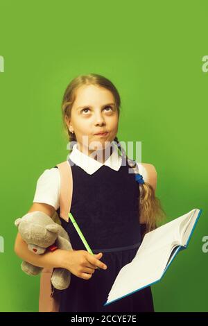 Kid in school uniform isolated on green background. Girl with braids and thoughtful face expression. Study and back to school concept. Pupil holds blue book, marker and teddy bear Stock Photo