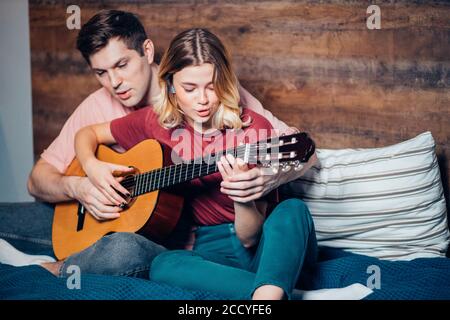 caucasian lovely couple play guitar together sitting on bed, young man teaching her girlfriend to play the guitar Stock Photo