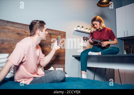 nice young caucasian female playing ukulele while his friend or boyfriend shot video or take photo of her, process of playing at home. leisure time Stock Photo