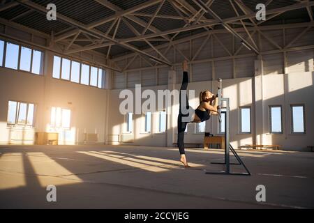 Charming young female acrobat practicing in gymnasium, doing splits in the air, standing on tiptoe holding ballet bar, professional sport concept Stock Photo
