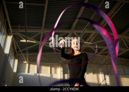 Close view of charming optimistic girl playing with violet ribbon, looking down with glad emotions, stretching hand towards camera, indoor shot from b Stock Photo