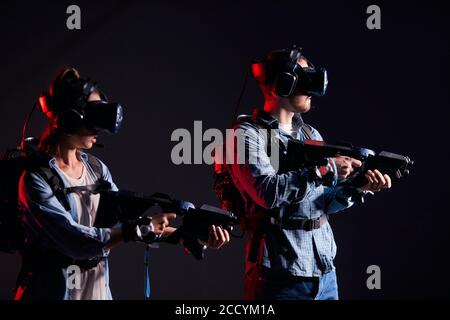 Couple of virtual reality gamers stand in neon backlit. Man and woman with virtual weapons isolated over black backgrond Stock Photo