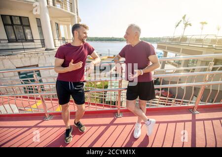 Yound male and grey-haired male leaning on bridge railing, resting during running Stock Photo