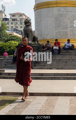 Young male monk asking for alms, food or money. Dressed in traditional red clothes, shaved head. Carrying alm bowl. Walking barefoot. Yangon, Myanmar Stock Photo
