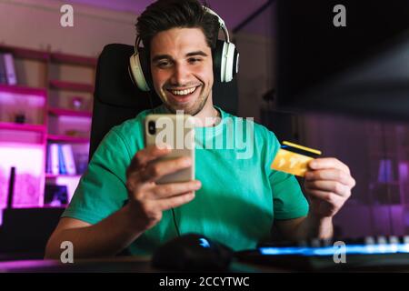 Image of caucasian smiling man holding credit card and cellphone playing video game on computer at home Stock Photo