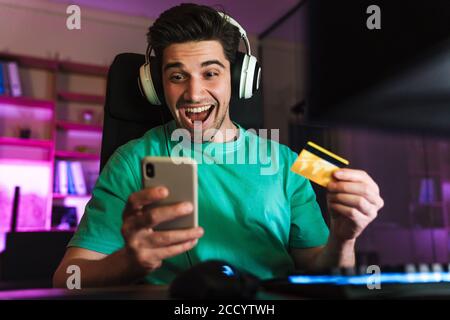 Image of young excited man holding credit card and cellphone playing video game on computer at home Stock Photo
